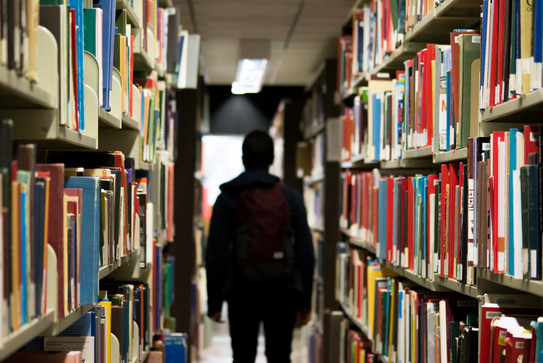 A male student stands in a library between two bookshelves 